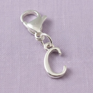 May include: A silver-toned metal charm in the shape of the uppercase letter C, featuring a smooth, reflective surface. The letter charm is attached to a functional silver-toned lobster claw clasp by a small, circular jump ring connector.