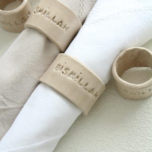 Könnte beinhalten: Nahaufnahme von Serviettenringen aus Keramik mit geprägtem Text. Die Ringe sind beige und umgeben gerollte weiße und beige Servietten. Der Text auf den Ringen lautet "Bismillah" und "Smillah".