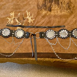 May include: A black belt with silver-colored embellishments and dangling chains. The belt features circular mirrored accents surrounded by clear crystals. A separate black strap and two silver rings are also included.