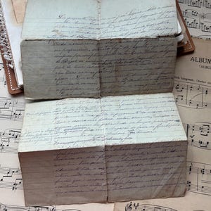 Könnte beinhalten: Vintage handgeschriebene Briefe und Ephemera, mit gealtertem Papier und verblasster Tinte. Die Briefe sind gefaltet und gestapelt, mit Noten im Hintergrund. Ein kleines blaues Buch mit dem Text "TOUS GENRES...DAIN BLEU" ist sichtbar.