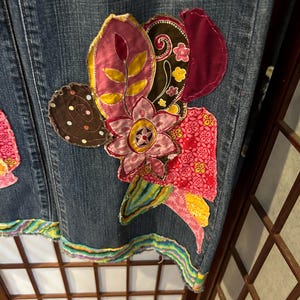 Op de afbeelding: Een denim rok met een kleurrijke bloemenapplicatie. De applicatie is gemaakt van verschillende stoffen en bevat de kleuren roze, rood, geel en bruin.