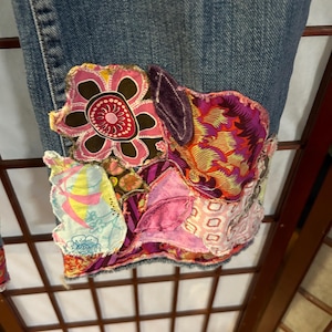 May include: Blue denim jeans with colourful patchwork detailing on the bottom hem. The patchwork includes floral, geometric, and abstract patterns in shades of pink, purple, yellow, and brown.