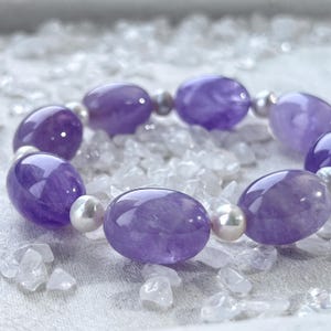 May include: A bracelet with large, oval, polished purple amethyst stones and small, round, white pearls. The bracelet is set on a bed of clear, crushed crystals, creating a sparkling effect.