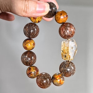 May include: A bracelet made of natural gemstones in shades of brown, orange, and gold. The bracelet features large, round beads and a clear, faceted stone. The bracelet is held up by a hand. The beads are approximately 2 cm in diameter.