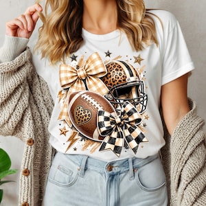 May include: A white t-shirt featuring a graphic print of a brown football with a leopard print heart, a leopard print football helmet, and two large bows in tan plaid and black and white checkered patterns, accented with gold stars and lightning bolts.