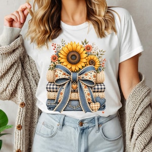 Könnte beinhalten: Ein weißes T-Shirt mit einem Grafikdruck eines Stapels gefalteter Stoffe, gekrönt von Sonnenblumen. Eine große, gemusterte blaue Schleife ist um den Stapel gebunden und mit kleinen herzförmigen Anhängern verziert. Das Design zeigt Denim- und Karotexturen.