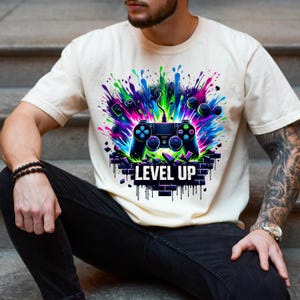 May include: Cream-colored t-shirt featuring a vibrant graphic of a video game controller with colorful paint splatter effects. The design includes the words "LEVEL UP" in bold white letters, perfect for gamers.