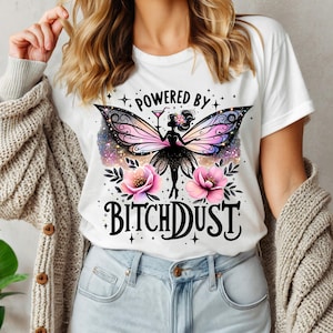 Op de afbeelding: Wit T-shirt met een afbeelding van een fee met kleurrijke vleugels, een cocktail vasthoudend, en de tekst "POWERED BY BITCHDUST". Het ontwerp bevat roze bloemen en sterren.