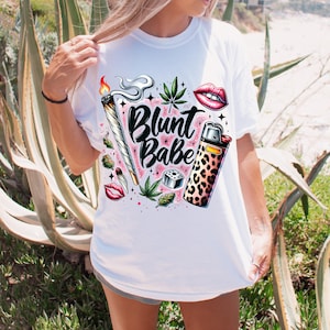 Peut inclure: T-shirt blanc avec un motif graphique coloré. Le motif comprend un joint allumé, des feuilles de cannabis, des lèvres rouges et un briquet à imprimé léopard. Les mots "Blunt Babe" sont écrits en cursive.