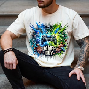 Puede incluir: Camiseta color crema con un diseño gráfico vibrante. El diseño presenta un mando de videojuegos negro en el centro, rodeado de explosiones azules y verdes, formas geométricas y el texto "GAMER BOY" en una fuente tipo ladrillo.