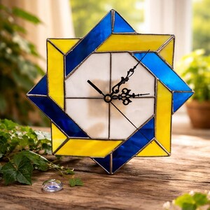 May include: A stained glass clock with a geometric design. The clock face is white, with black hands. The frame is a combination of yellow and blue glass, creating a star-like shape. The clock sits on a wooden surface.