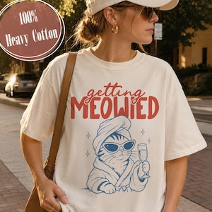 Op de afbeelding: Een off-white t-shirt met korte mouwen en de rode tekst "getting MEOWIED" boven een blauwe afbeelding van een kat met een handdoek op het hoofd, een zonnebril en een badjas, terwijl het dier een champagneglas vasthoudt.