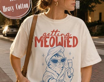 May include: An off-white short-sleeve t-shirt featuring the red text "getting MEOWIED" above a blue graphic of a cat wearing a towel on its head, sunglasses, and a bathrobe while holding a champagne flute. A circular badge reads 100% Heavy Cotton.