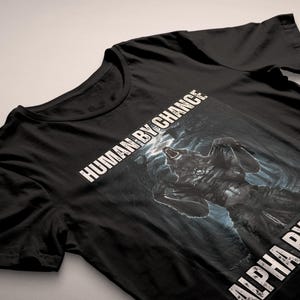Puede incluir: Camiseta negra con un gr&aacute;fico de lobo y el texto "HUMAN BY CHANCE" y "ALPHA BY CHOICE" en blanco. El lobo se muestra en un entorno oscuro y dram&aacute;tico, con una expresi&oacute;n de aullido.