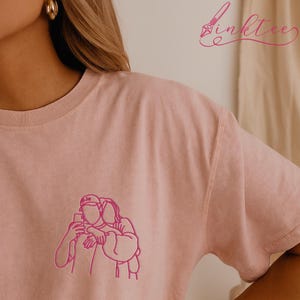 May include: A light pink t-shirt featuring a pink line drawing of two people embracing while taking a selfie. The shirt has a crew neck and short sleeves. The brand name "inktee" is written in pink script in the upper right corner.