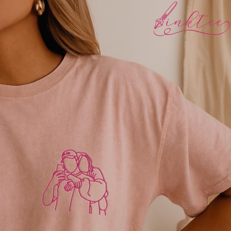 May include: A light pink t-shirt featuring a pink line drawing of two people embracing while taking a selfie. The shirt has a crew neck and short sleeves. The brand name "inktee" is written in pink script in the upper right corner.