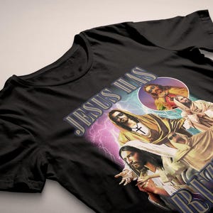 May include: Black t-shirt featuring a graphic design with the text "JESUS HAS RIZZEN" and images of Jesus. The design includes a cross, lightning bolts, and a circular image.