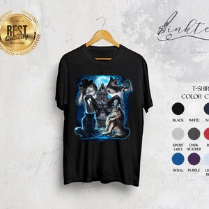 May include: Black t-shirt with a fantasy illustration of werewolves under a full moon. The graphic includes a central wolf figure and other wolf-like creatures. The shirt is displayed on a wooden hanger against a neutral background. Color chart included.