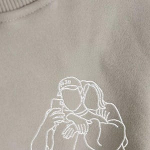May include: A beige sweatshirt with a white embroidered outline of two people taking a selfie. One person is wearing a baseball cap. The sweatshirt has a crew neck and a soft, textured appearance.