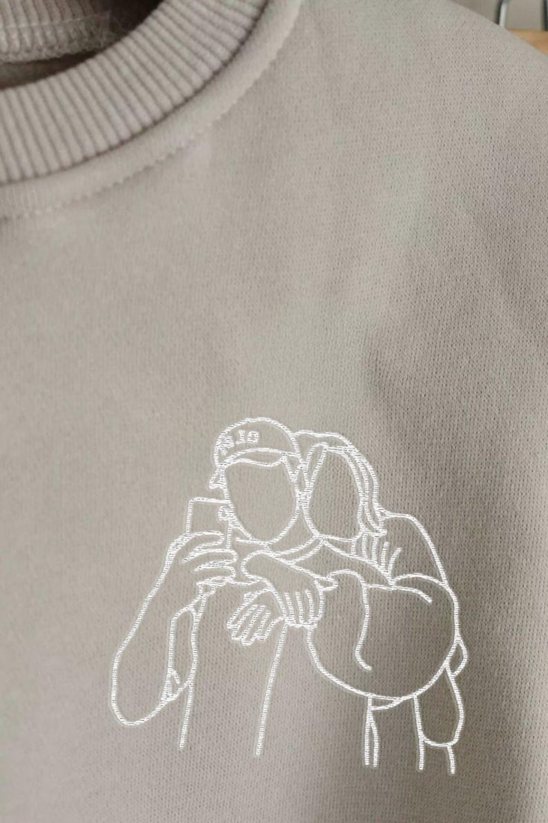 May include: A beige sweatshirt with a white embroidered outline of two people taking a selfie. One person is wearing a baseball cap. The sweatshirt has a crew neck and a soft, textured appearance.