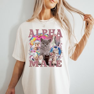 May include: A white t-shirt with a pink graphic that says "ALPHA MALE" and features a collage of cats and kittens. The graphic includes a rainbow, a unicorn, and a star.