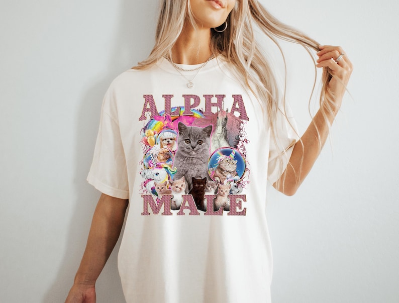 May include: A white t-shirt with a pink graphic that says "ALPHA MALE" and features a collage of cats and kittens. The graphic includes a rainbow, a unicorn, and a star.