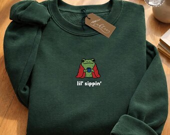 May include: Dark green sweatshirt with an embroidered frog design. The frog is wrapped in a red blanket and holding a blue cup. The text "lil' sippin'" is below the frog. A brown tag is attached to the collar.