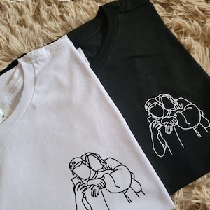 May include: Two t-shirts, one white and one black, are displayed. Each shirt features a minimalist line drawing of two people embracing while looking at a phone. The shirts are on a textured, light brown surface.