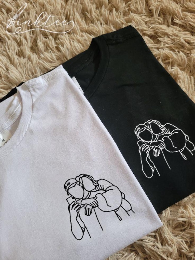 May include: Two t-shirts, one white and one black, are displayed. Each shirt features a minimalist line drawing of two people embracing while looking at a phone. The shirts are on a textured, light brown surface.