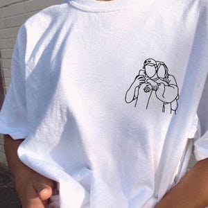 May include: White t-shirt with a small black line drawing of two people taking a selfie. The shirt has a crew neck and short sleeves. The brand name "inktee" is written in cursive in the upper left corner.