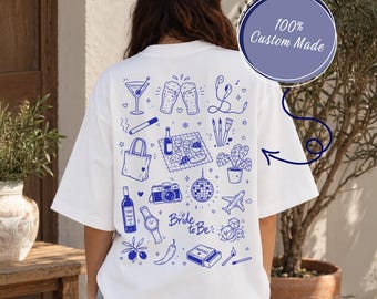 May include: A white t-shirt featuring a blue graphic print on the back with various icons including a martini glass, beer mugs, headphones, a tote bag, a picnic scene, a camera, a disco ball, an airplane, and the text Bride to Be.