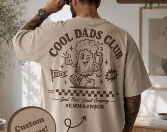 May include: A sand-colored t-shirt featuring a dark brown graphic on the back. The design includes a cartoon beer mug, the text COOL DADS CLUB, ESTD 2026, and the phrase Good Beer Great Company. Below this, the custom names #EMMA and #NICK are printed.
