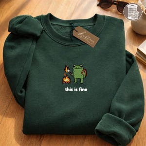 May include: A dark green sweatshirt with an embroidered frog design. The frog, wearing a backpack, is roasting a marshmallow over a campfire. The text "this is fine" is below the design. A brown tag is attached to the neckline.