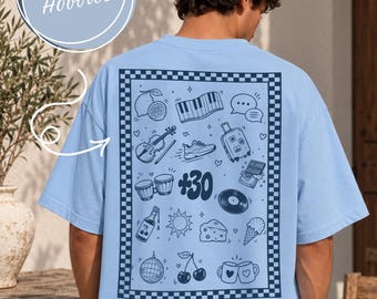 May include: A light blue t-shirt featuring a dark blue rectangular checkerboard border on the back. Inside the border are various line art illustrations including a piano, violin, suitcase, sneakers, cherries, disco ball, and the text +30.