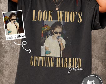 May include: A dark heather gray short-sleeve t-shirt featuring a custom photo print of a child wearing sunglasses and holding a microphone. The text "LOOK WHO'S GETTING MARRIED" appears in gold lettering above and below the photo, with the name "Julia" in white script.