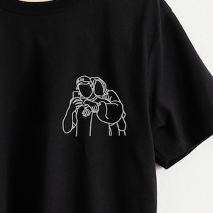 May include: Black t-shirt featuring a white line drawing of a couple taking a selfie. The design is on the left chest area. The shirt has short sleeves and a crew neck. The brand name "inktee" is written in cursive on the top right.