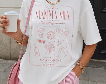 May include: A white short-sleeve t-shirt featuring a pink graphic print with the text MAMMA MIA, I'm getting married, BRIDE TO BE, SANTORINI, and SEPT 20, 2026. The design includes illustrations of a disco ball, champagne, and flowers.