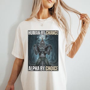 Puede incluir: Camiseta color crema con un gr&aacute;fico de un hombre lobo transform&aacute;ndose. La imagen est&aacute; ambientada sobre un fondo oscuro y cavernoso. El texto de la camiseta dice "HUMAN BY CHANCE" encima de la imagen y "ALPHA BY CHOICE" debajo.