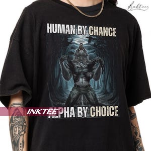 Puede incluir: Camiseta negra con un dise&ntilde;o de hombre lobo transform&aacute;ndose, sobre un fondo de bosque oscuro. El texto "HUMAN BY CHANCE" est&aacute; encima de la imagen y "ALPHA BY CHOICE" debajo. La camiseta tiene un corte holgado.