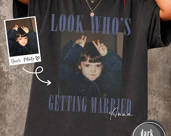 May include: A dark heather gray short-sleeve t-shirt featuring a custom photo print of a child making a peace sign gesture. The text "LOOK WHO'S GETTING MARRIED" appears in blue above and below the photo, with the name "Anna" in white script.