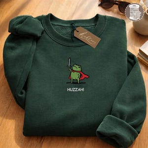 Puede incluir: Sudadera verde oscuro con un bordado de una rana con capa roja y espada. La palabra "Huzzah!" está bordada debajo. Una etiqueta marrón con texto en cursiva cuelga del cuello.