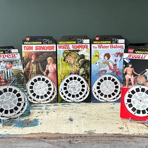 Könnte beinhalten: Eine Sammlung von Vintage View-Master-Rollen und -Verpackungen. Zu den Titeln gehören "Flipper", "Tom Sawyer", "Worzel Gummidge", "The Water Babies" und "Woogly". Jede Rolle ist weiß mit einem schwarzen Design, und die Verpackung zeigt farbenfrohe Illustrationen.