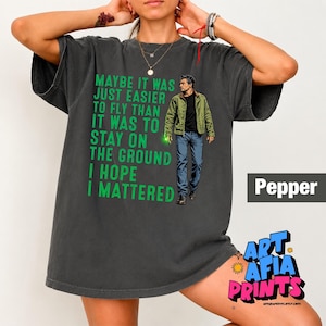 May include: A charcoal gray short-sleeve t-shirt featuring a graphic of a man in a green jacket and blue jeans. The text reads: MAYBE IT WAS JUST EASIER TO FLY THAN IT WAS TO STAY ON THE GROUND I HOPE I MATTERED. The shirt has a crew neckline.