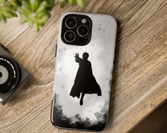 May include: A smartphone case with a black silhouette of a person in a cape, set against a white and gray watercolor-style background. The case has a black border and a camera cutout. The phone case is on a wooden surface.