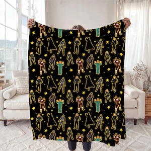 Op de afbeelding: Zwarte deken met een herhalend patroon van figuren in verschillende outfits, waaronder capes en uniformen, en gele sterren. De figuren zijn geel omlijnd. De deken wordt voor een raam en een witte bank gehouden.