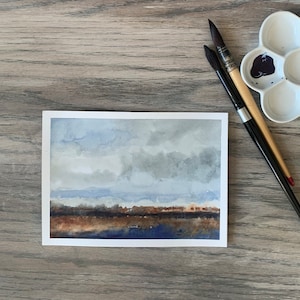 May include: A watercolor painting of a landscape with a cloudy sky, a line of trees, and a body of water. Two paintbrushes and a white paint palette with paint wells are on a wooden surface.