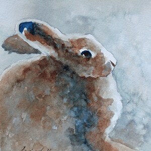 K&ouml;nnte beinhalten: Aquarellmalerei eines braunen Kaninchens mit blauen Augen, das auf einem hellblauen Hintergrund sitzt. Das Kaninchen schaut nach rechts. Die Unterschrift des K&uuml;nstlers befindet sich in der linken unteren Ecke.