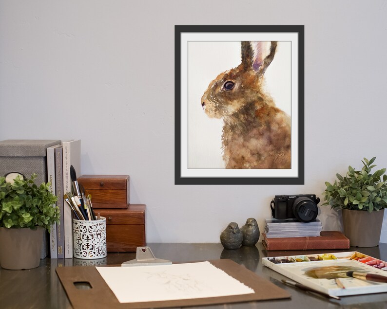 May include: Watercolor painting of a brown rabbit with long ears, looking to the left. The rabbit is painted in shades of brown, with a white background.