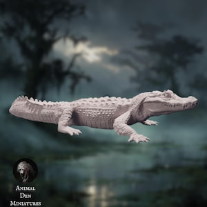 May include: A light-coloured alligator miniature, with detailed scales and a textured body, is displayed against a blurred swamp background. The miniature is a detailed representation of the reptile. The text "Animal Den Miniatures" is visible in the lower left corner.