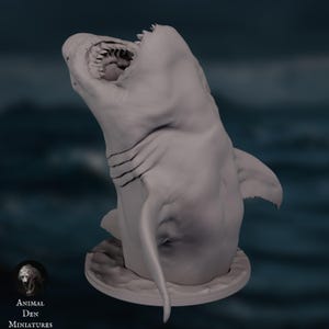 May include: A gray, three-dimensional sculpture of a shark with its mouth open, revealing sharp teeth. The shark is positioned at an angle, showcasing its dorsal fin and body details. The sculpture is on a circular base, with the Animal Den Miniatures logo in the lower left corner.